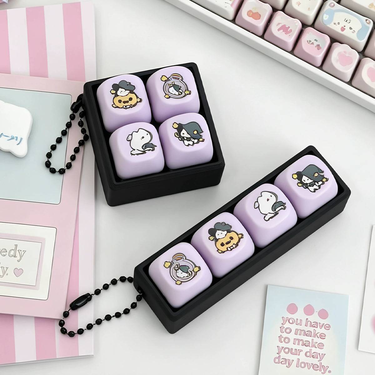 Magic cat keycap clicker keychains with lavender keycaps in black cases on a pink desk